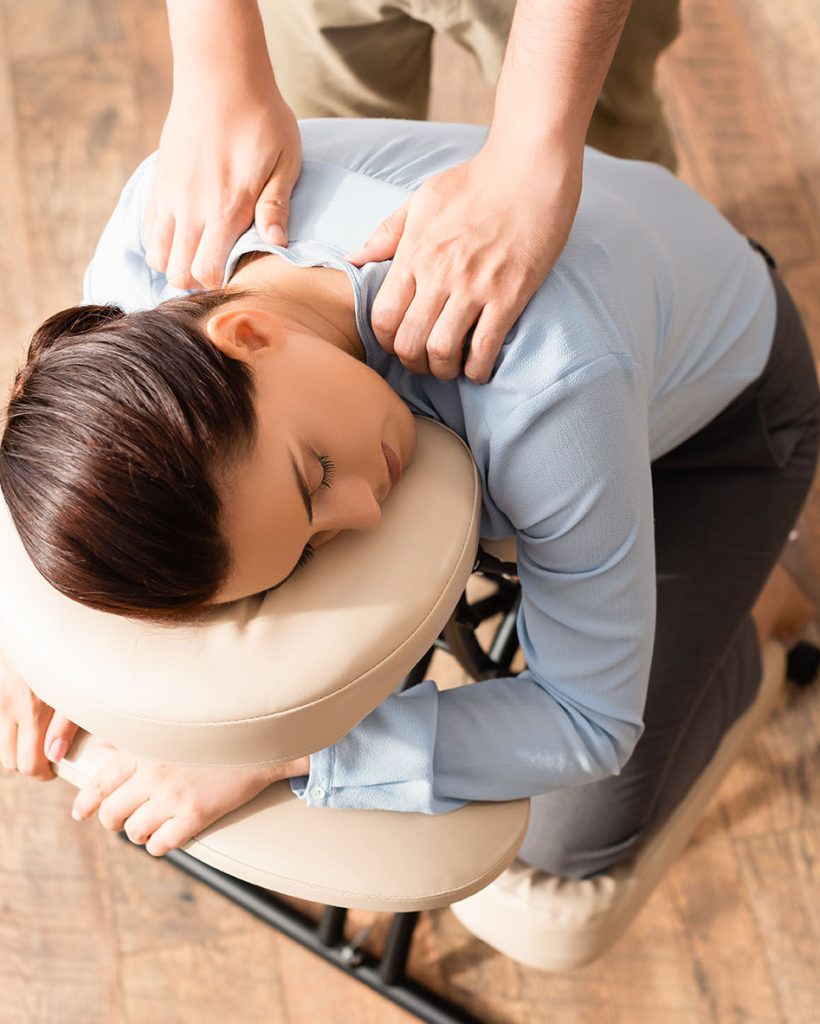 Overhead view of masseur massaging shoulders of brunette businesswoman with closed eyes, sitting on massage chair